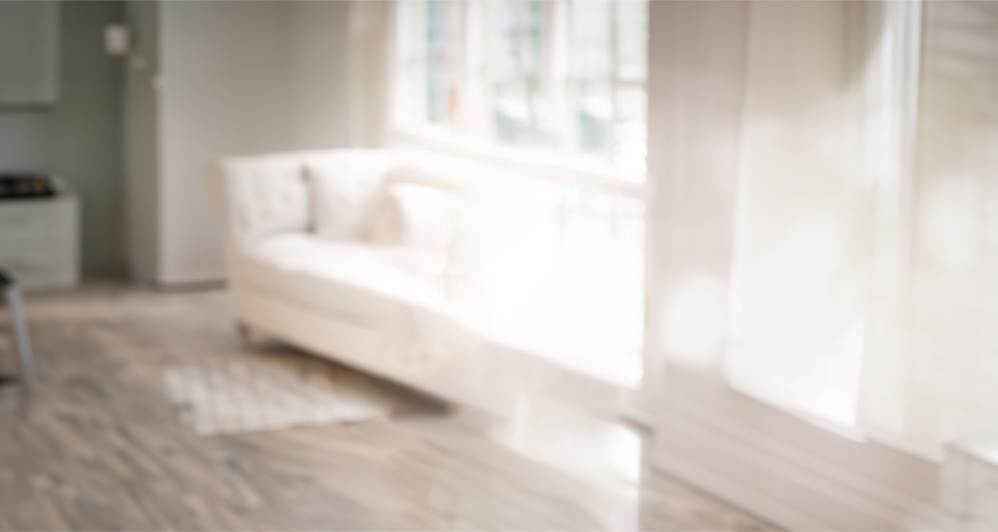 A bright and blurry image of a living room. The room has large windows allowing natural light to stream in. There is a white sofa placed near the windows, and the floor appears to be wooden. The overall effect is hazy, giving an ethereal and soft focus feel to the space.